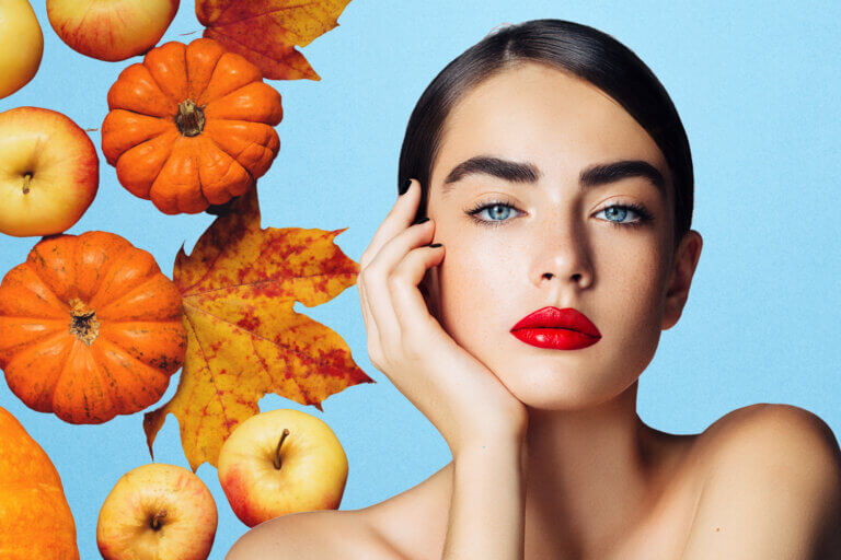 A model with dark hair and red lips. Apples, pumpkins and fall leaves behind her.